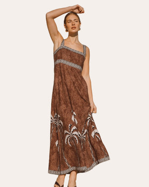 Woman in a brown dress with patterns standing on a sandy path with greenery in the background